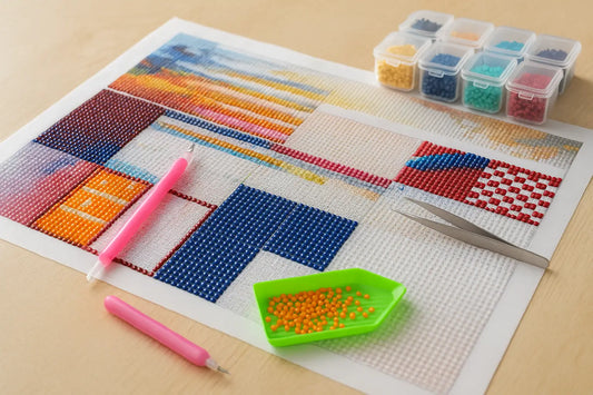 Onvoltooide diamond painting met kleurrijke vakken blauw, rood, oranje en geel, naast roze pennen, pincet, groene bakje met oranje steentjes en opbergdoos met kralen.