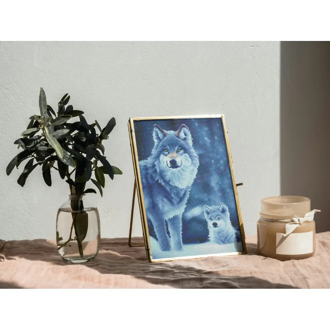 Diamond painting van een wolf met een welpje in een winterlandschap, geplaatst op een oppervlak naast een glazen vaas met een plant en een kaars.