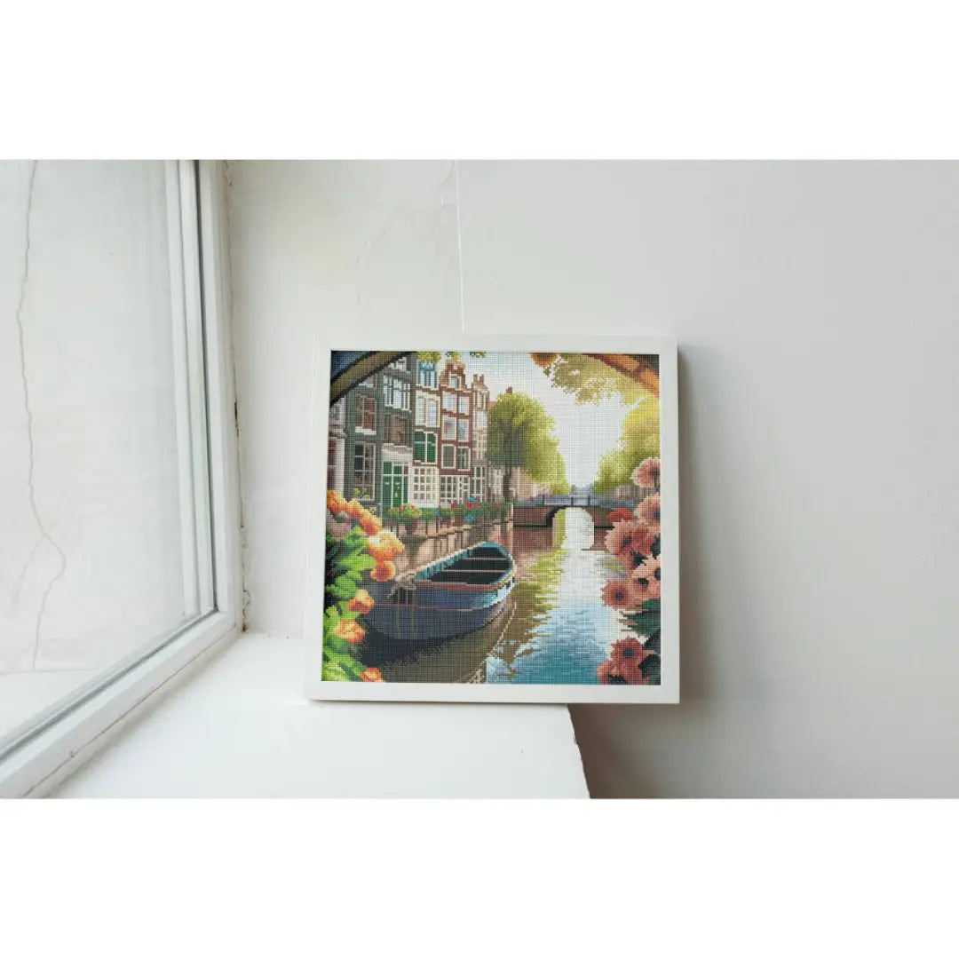 Ingelijste diamond painting van een sfeervol Amsterdams grachtenpand met brug, boot en bloemen langs het water in zacht zonlicht, geplaatst op een vensterbank.